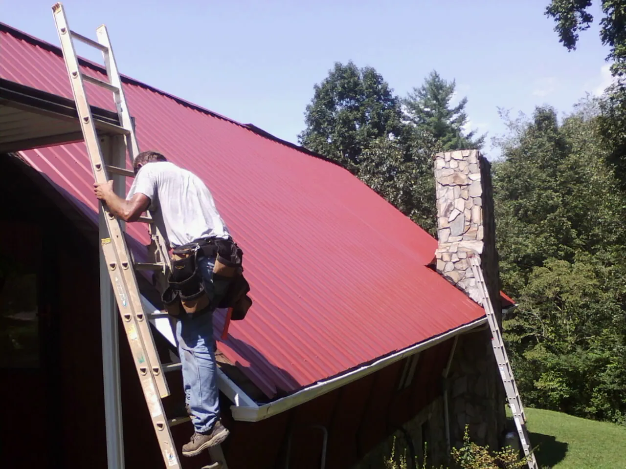 Roofer on a ladder beside a red metal roof and stone chimney — Soffit & Fascia Repair by Roofing Brothers in Wimberley